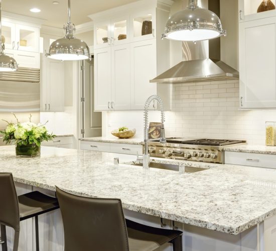White kitchen design features large bar style kitchen island with granite countertop illuminated by modern pendant lights. Stainless steel appliances framed by white shaker cabinets . Northwest, USA